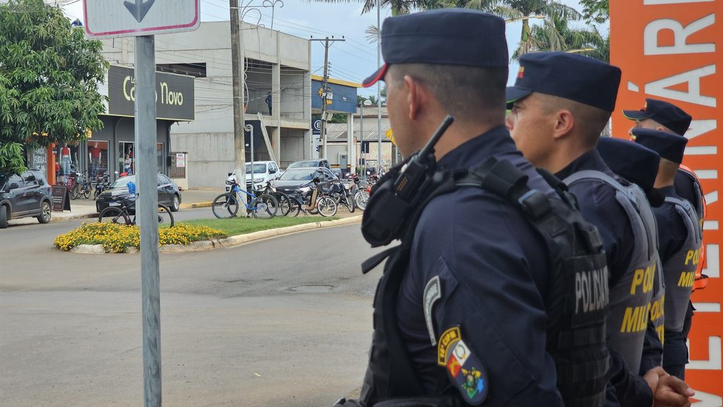 Alunos soldados da Polícia Militar fardados e com colete, em fila na calçada do Centro de Campo Novo do Parecis, com canteiro florido e lojas ao fundo