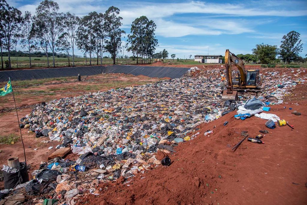 Retroescavadeira espalha lixo doméstico em célula de aterro sanitário com manta impermeável ao fundo