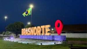 Letreiro iluminado de Brasnorte à noite, com bandeira do Brasil ao fundo e marcador de localização vermelho
