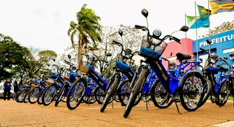 Fileira de bicicletas elétricas azuis com cestinha estacionadas em frente à Prefeitura de Sapezal