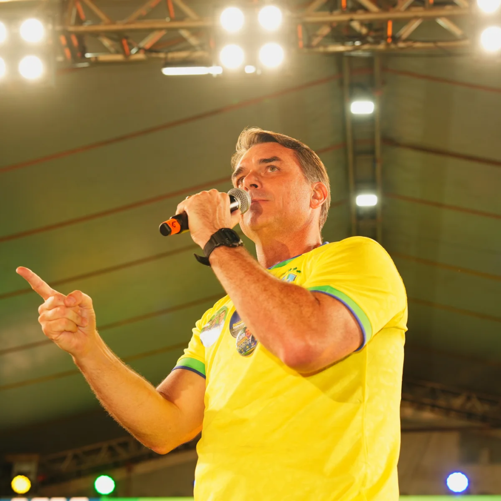 Flávio Bolsonaro, de camisa amarela, fala ao microfone e aponta com a mão em cima de um palco iluminado