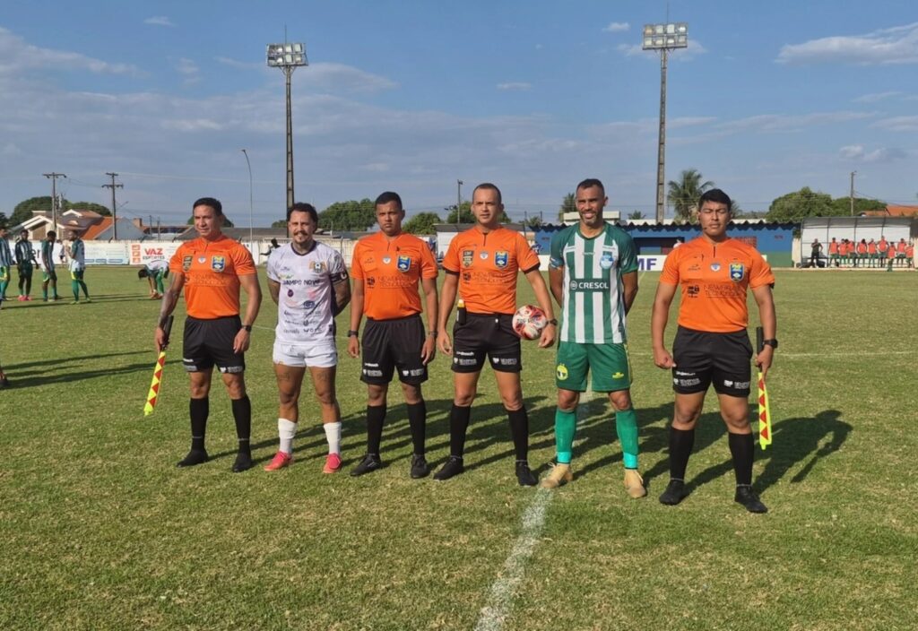 Quatro árbitros de uniforme laranja posados no gramado do Estádio Ary Tomazelli ao lado dos capitães do Campo Novo, de camisa branca, e do Sorriso, de camisa listrada verde e branca
