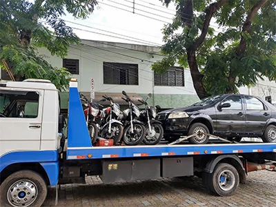 Caminhão guincho de plataforma azul transportando motos e um carro escuro recolhidos