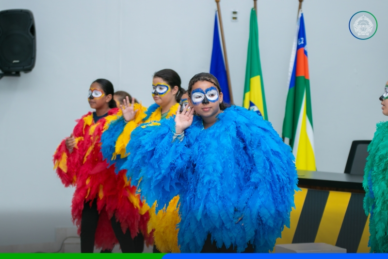 Alunos fantasiados de araras azul, vermelha e amarela acenam durante apresentação na Semana do Trânsito em Sapezal