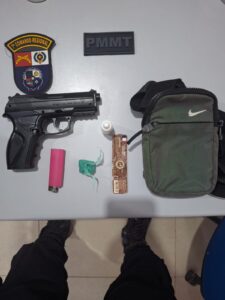Simulacro de pistola, porção de maconha, isqueiro, seda e bolsa transversal apreendidos com adolescente em escola, sobre mesa com emblemas da PMMT