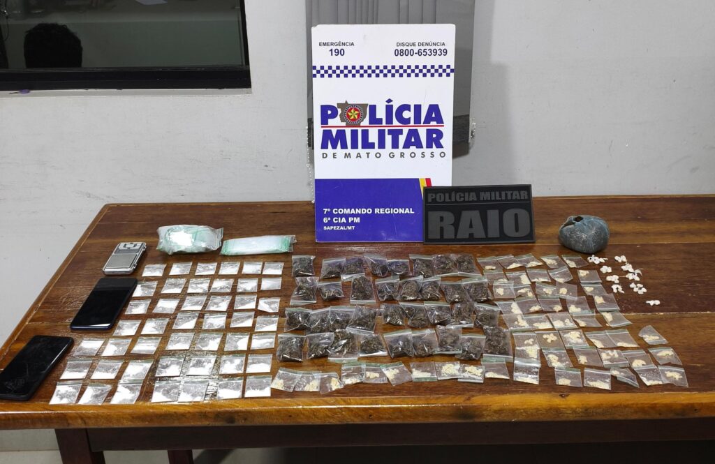 Porções de maconha, pasta base e cocaína, balança de precisão e celulares apreendidos sobre mesa, com placas da Polícia Militar e da equipe Raio