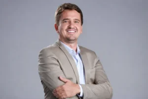 Vereador Norberto Junior, de Brasnorte, de paletó bege e camisa azul-clara, sorrindo de braços cruzados diante de fundo cinza