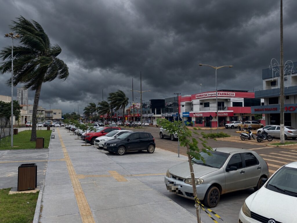 Nuvens escuras sobre avenida de Campo Novo do Parecis, com coqueiros balançando com o vento e carros estacionados