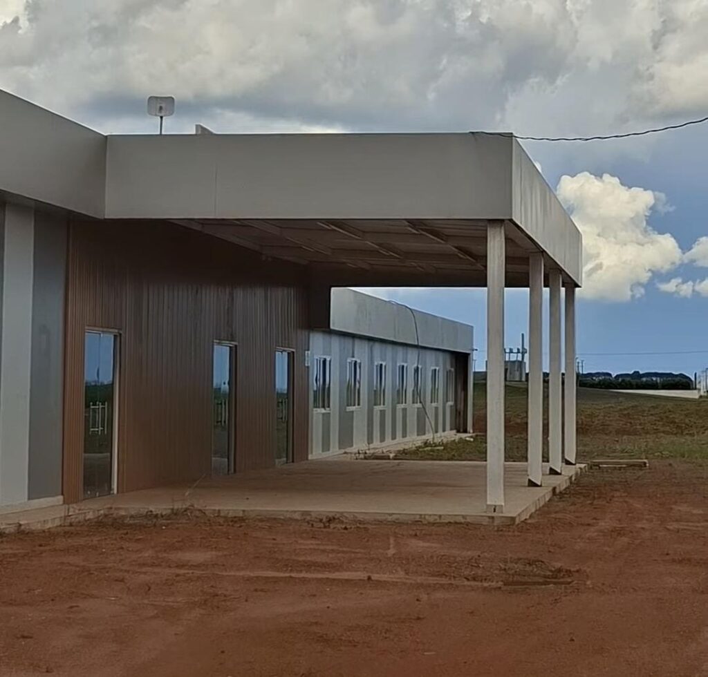 Prédio da faculdade de Sapezal, com fachada de placas metálicas, janelas de vidro e cobertura na entrada, cercado por terra vermelha