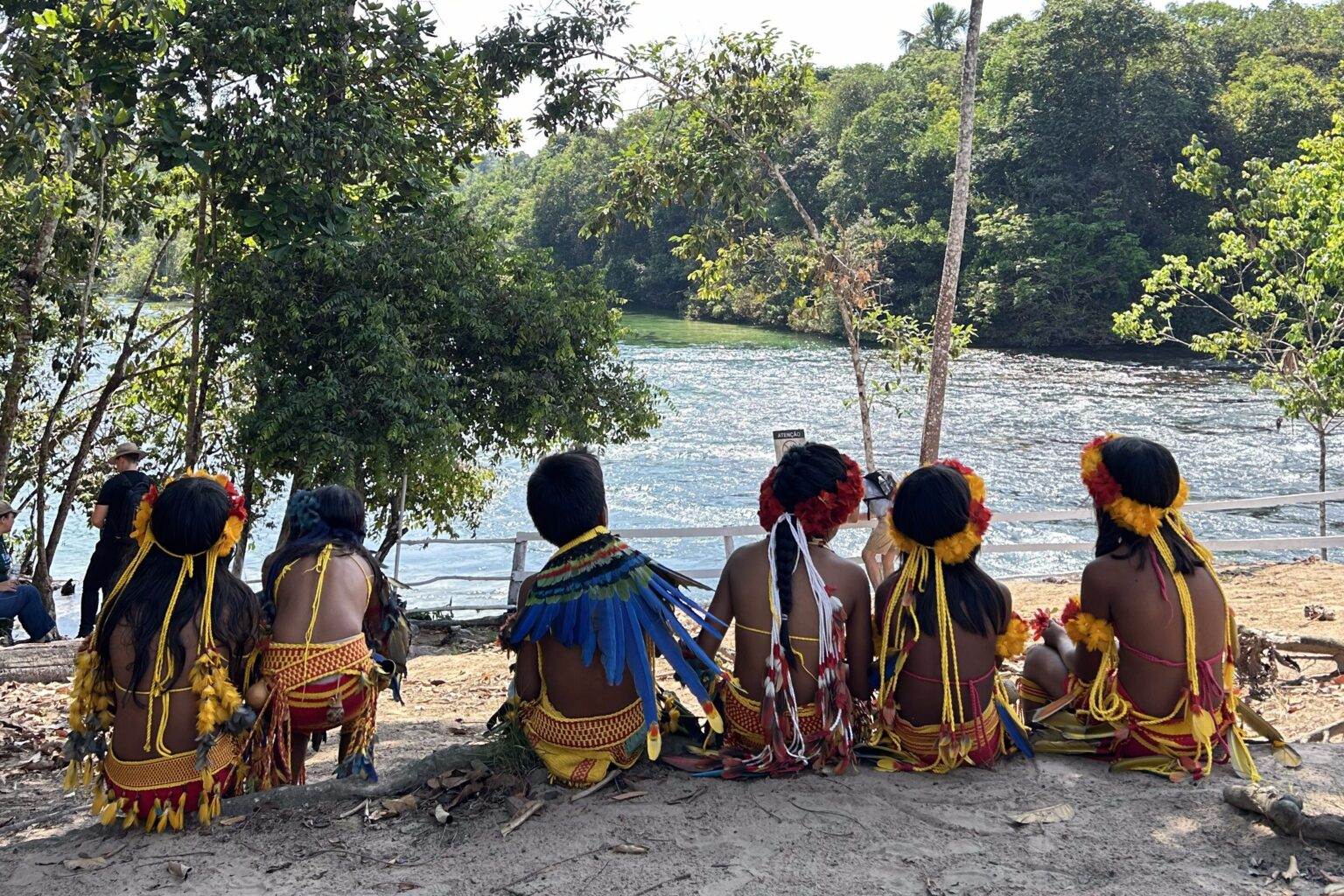Crianças indígenas com adornos de penas e cordões amarelos, vistas de costas, sentadas diante de um rio cercado de mata