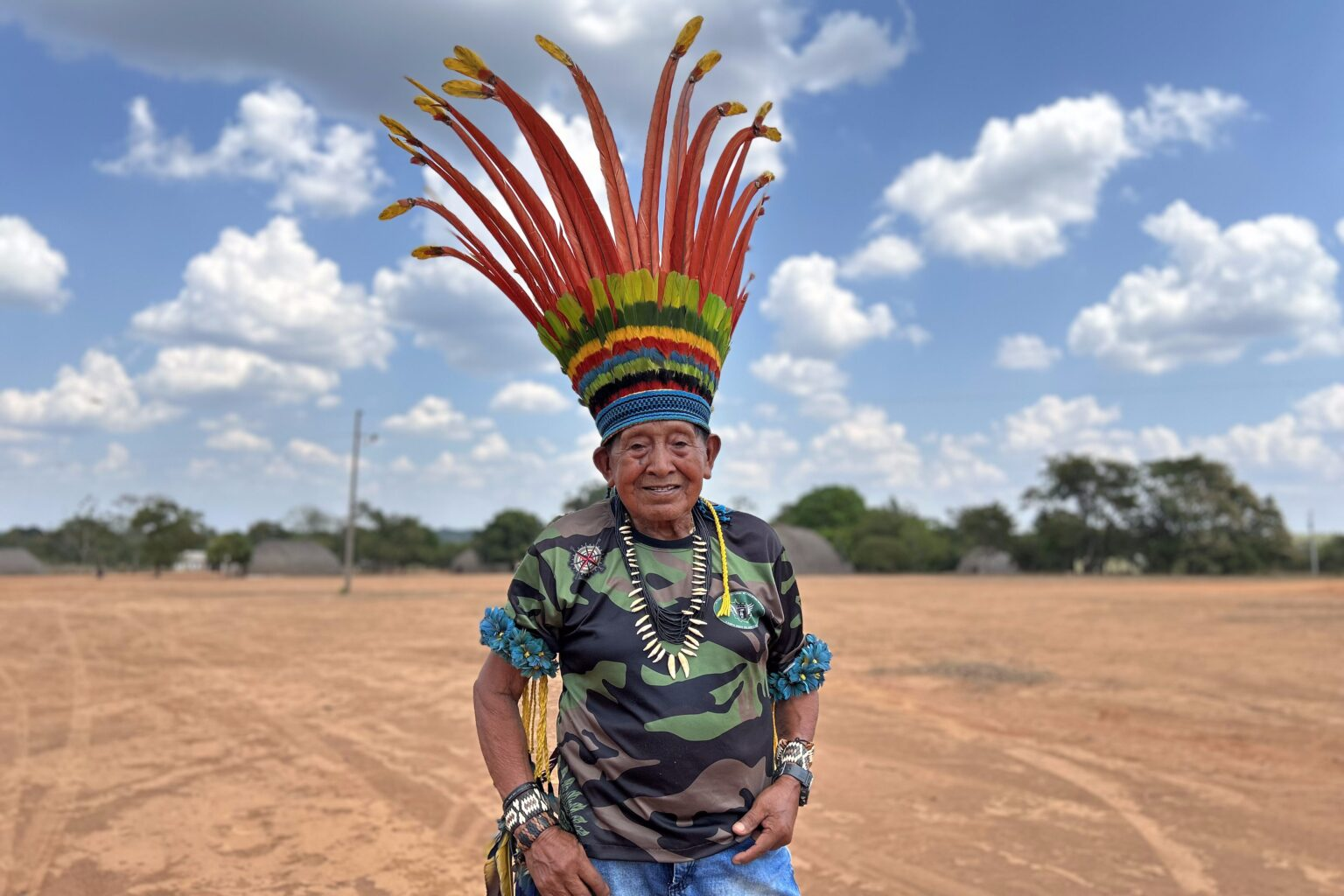 Cacique idoso com grande cocar de penas vermelhas e coloridas, colar de dentes e camiseta camuflada, em pátio de terra da aldeia