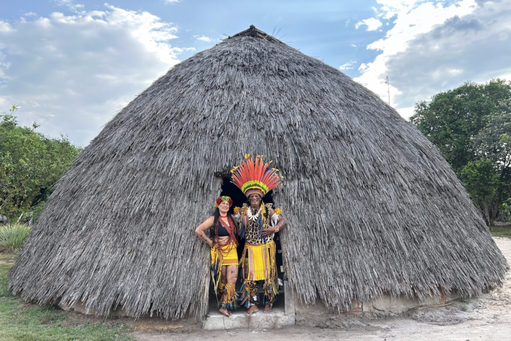 Casal indígena Haliti-Paresi com trajes tradicionais e cocar de penas coloridas na porta de uma grande oca de palha na Aldeia Wazare