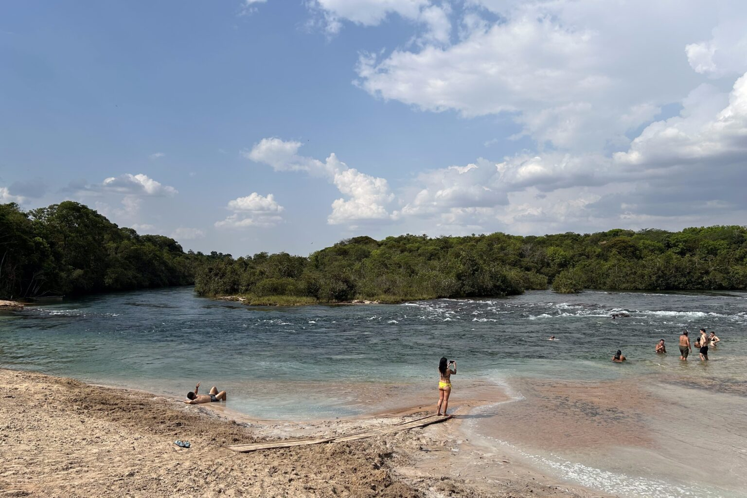 Visitantes tomam banho e tiram fotos em rio de águas claras com corredeiras e mata nas margens, na Aldeia Wazare