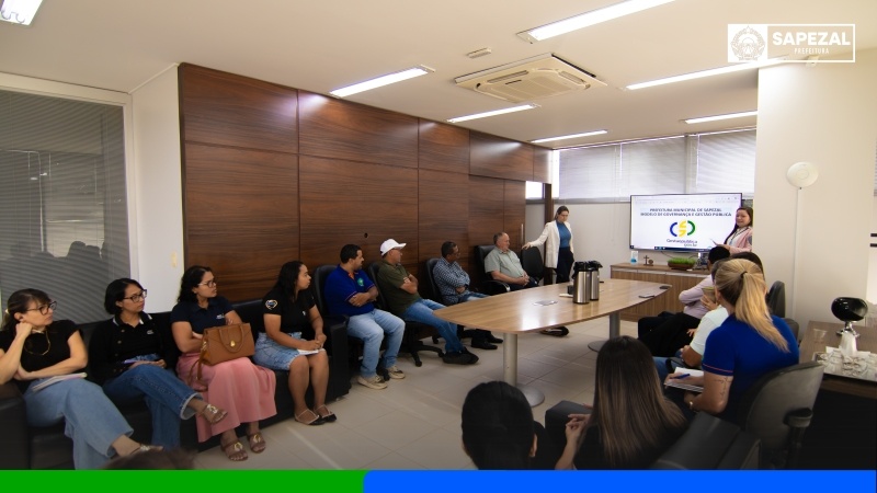Servidores sentados em sala de reunião da Prefeitura de Sapezal acompanham apresentação de uma mulher em pé ao lado de uma TV com o slide do programa de governança