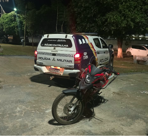 Moto vermelha e preta estacionada atrás de uma viatura da Polícia Militar, à noite, em uma área pavimentada com árvores ao fundo, em Brasnorte