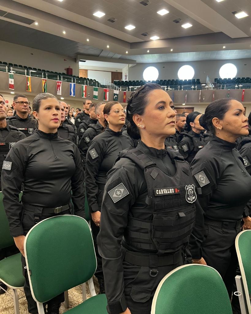 Policiais penais de farda preta perfilados em auditório durante solenidade, com bandeiras ao fundo