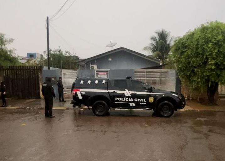 Viatura da Polícia Civil estacionada em frente a uma casa, com policiais na calçada, em dia de chuva