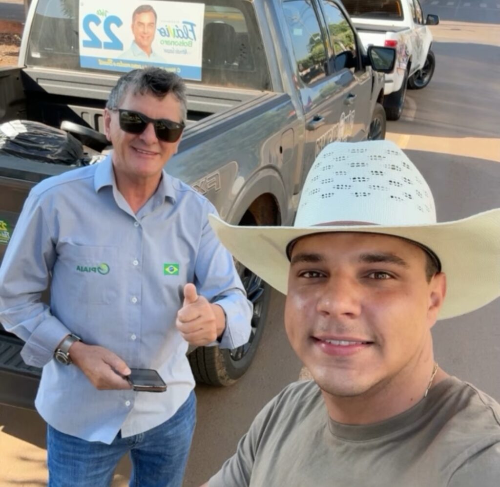 Prefeito Edilson Piaia, de óculos escuros e camisa azul-clara, faz sinal de positivo ao lado do vereador Djonathan Baioto, de chapéu branco, em frente a uma caminhonete com adesivo de Flávio Bolsonaro