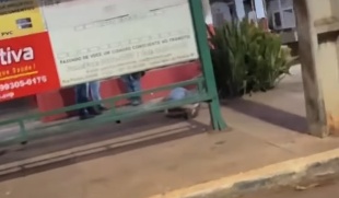 Imagem de vídeo, desfocada, mostra ponto de ônibus com placa de propaganda e pessoas na calçada à noite
