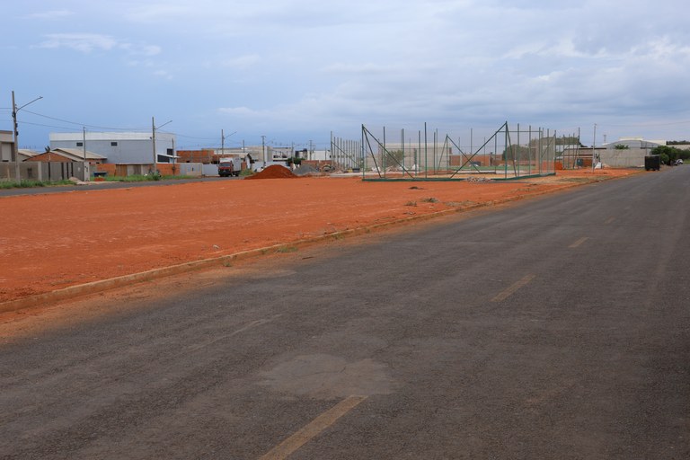 Rua asfaltada ao lado de terreno de terra vermelha com estrutura de quadra cercada por alambrado, no bairro Parque dos Girassóis, em Campo Novo do Parecis