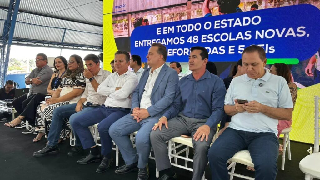 Autoridades sentadas em evento de entrega de escola, com painel sobre novas escolas estaduais ao fundo