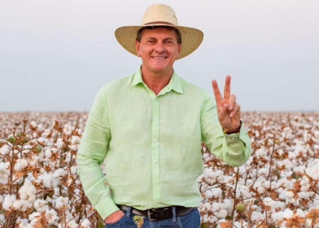 Prefeito Edilson Piaia, de chapéu de palha e camisa verde-clara, sorri e faz sinal de V com a mão em uma lavoura de algodão