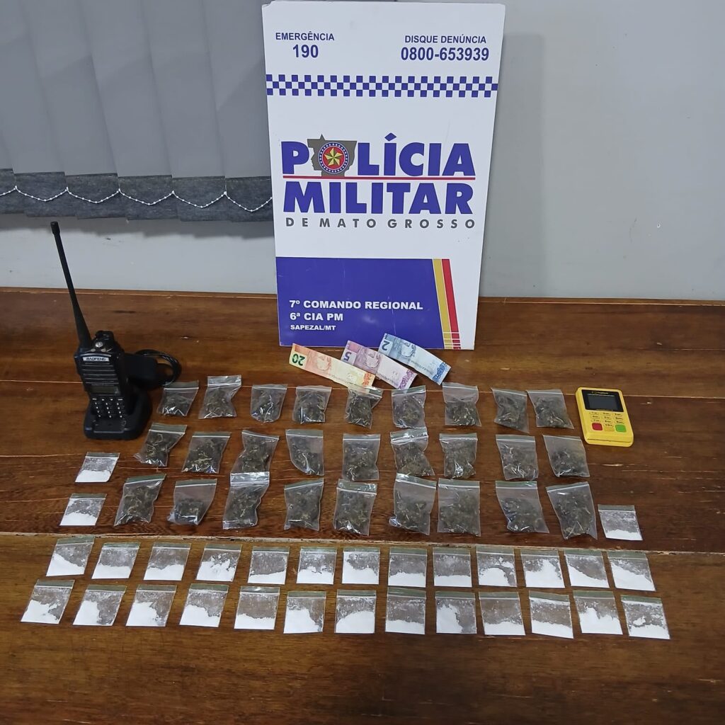 Porções de cocaína e maconha em saquinhos, rádio comunicador, máquina de cartão e notas de dinheiro sobre uma mesa, diante de placa da Polícia Militar