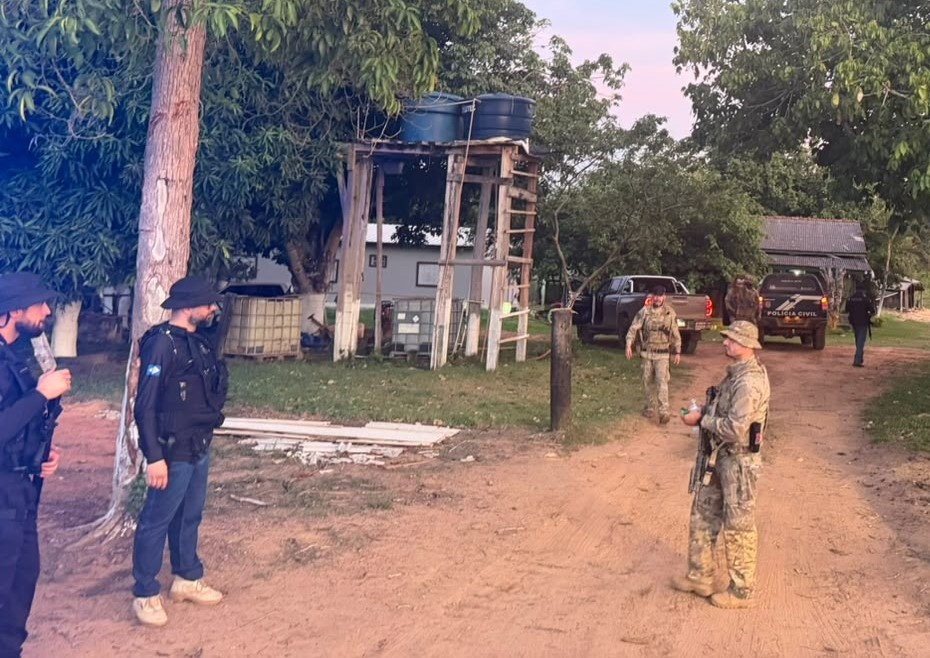 Policiais civis armados e fardados em uma propriedade rural, com caminhonetes e viatura da Polícia Civil ao fundo, no fim da tarde