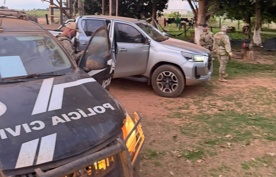 Viatura da Polícia Civil em primeiro plano e caminhonete prata com a porta aberta, cercada por policiais fardados em uma propriedade rural
