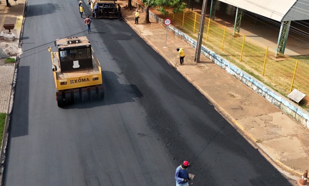 Vista aérea de rua recebendo asfalto novo, com rolo compactador amarelo, vibroacabadora e trabalhadores de uniforme amarelo