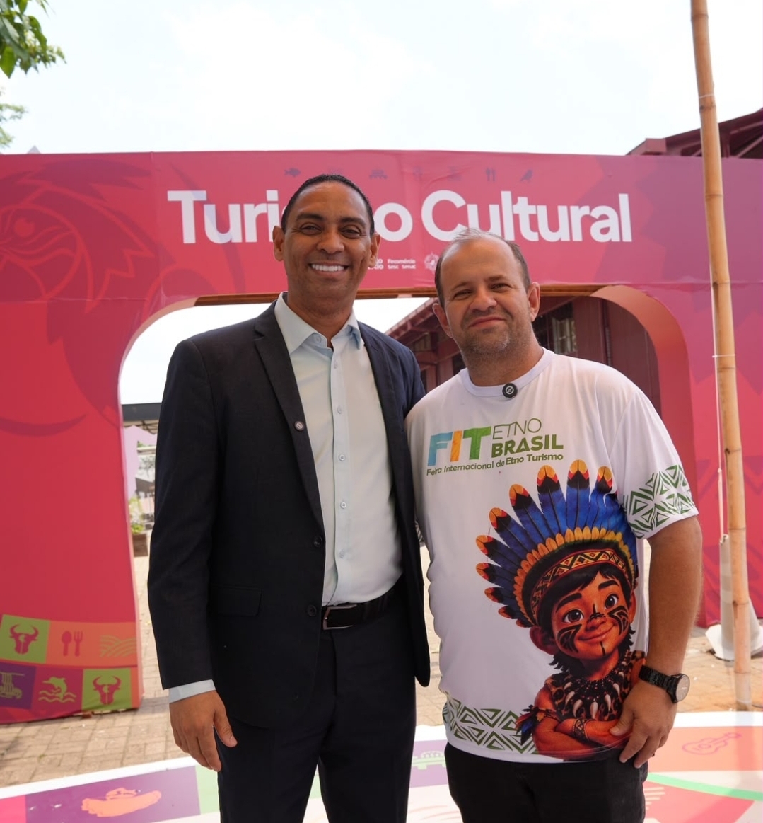 Secretário de Turismo de Campo Novo do Parecis, com camiseta da FIT Etno Brasil, posa ao lado de homem de terno em frente ao portal Turismo Cultural da Expo Turismo, em Porto Velho