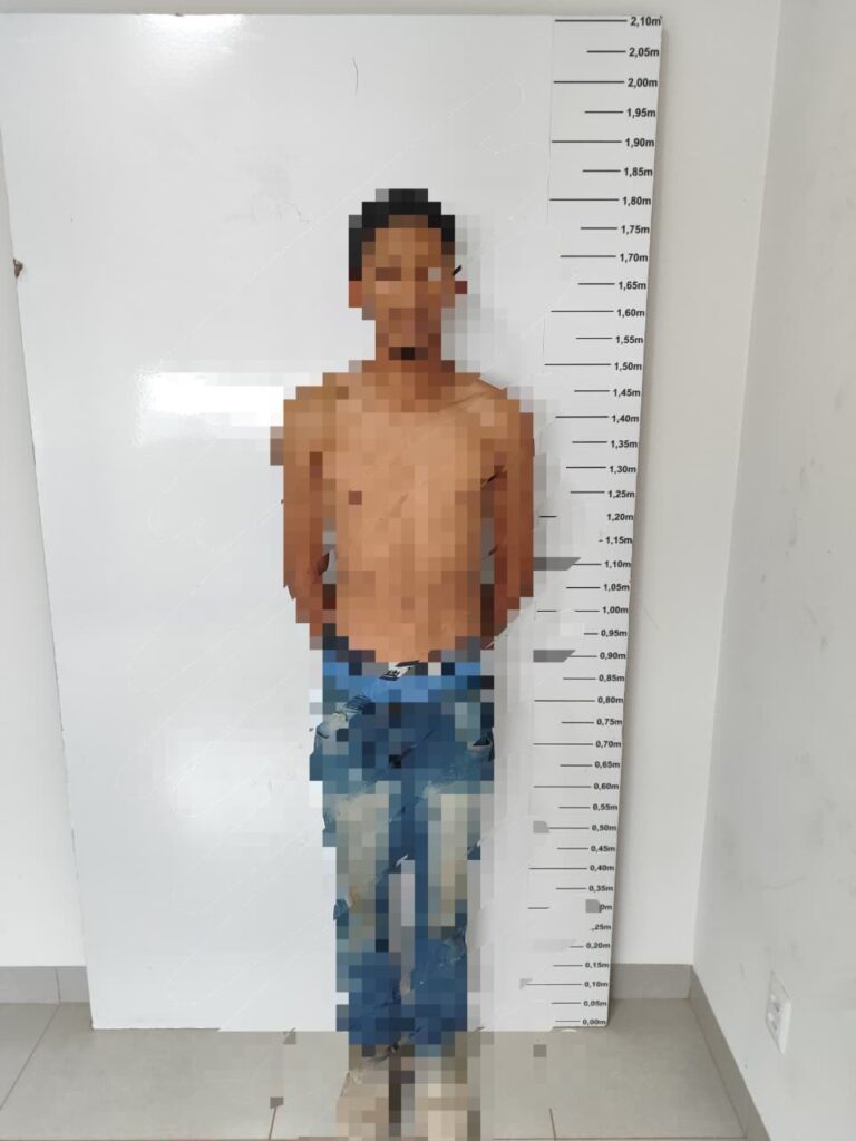 Homem sem camisa e de calça jeans, com o corpo e o rosto borrados, em frente a uma régua de altura na delegacia