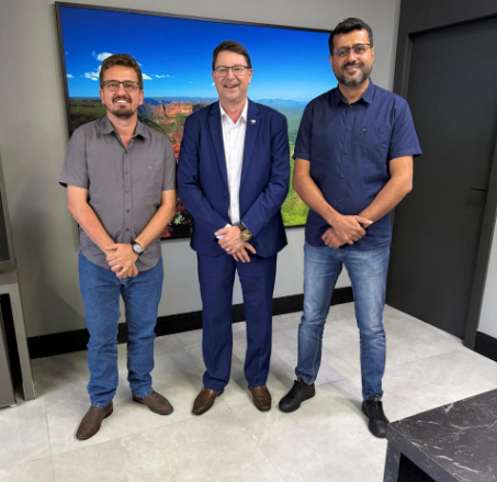 Três homens posam em sala de escritório, em frente a uma TV com paisagem, durante visita de vereadores de Brasnorte à Sinfra