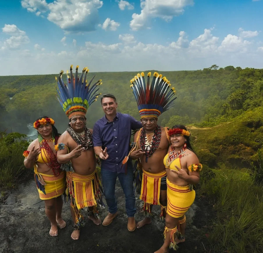 Flávio Bolsonaro posa ao lado de quatro indígenas Haliti-Paresi com cocares e trajes tradicionais, em mirante com cachoeira e mata ao fundo